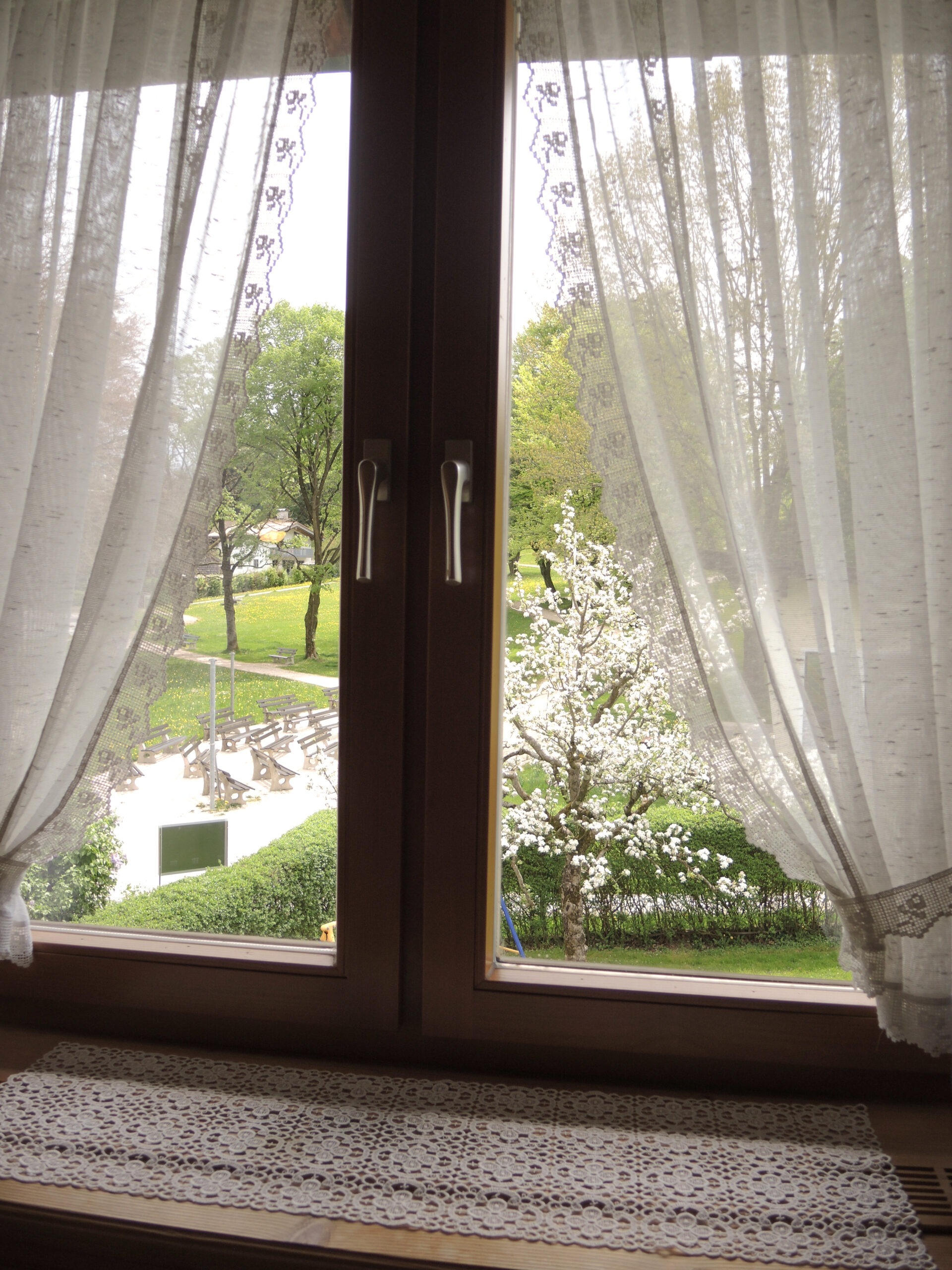 Der Blick aus einem Fenster, gerahmt von weißen Gardinen mit Spitze, auf blühende Bäume, grüne Wiesen und gepflegte Spazierwege im Sonnenschein.