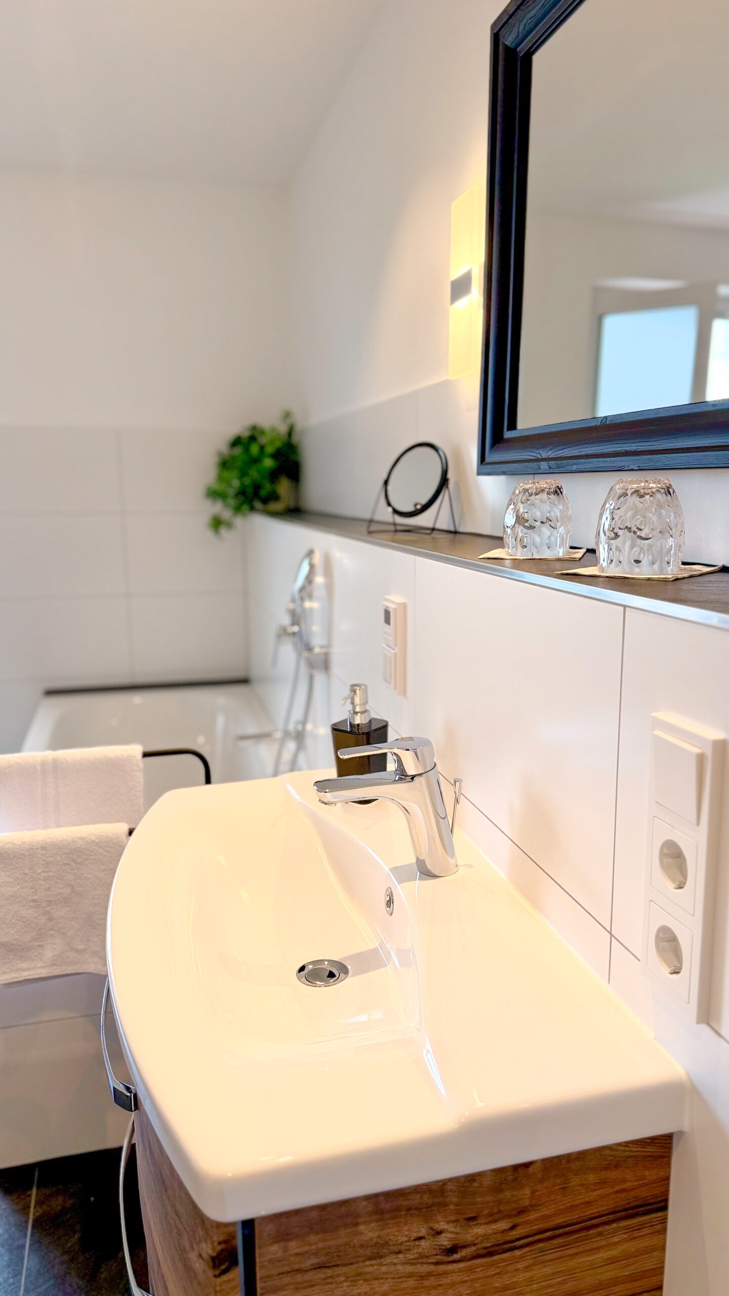 Ein modernes Badezimmer mit einem großen, geschwungenen weißen Waschbecken, einem Spiegel mit schwarzem Rahmen und frischen Trinkgläsern.