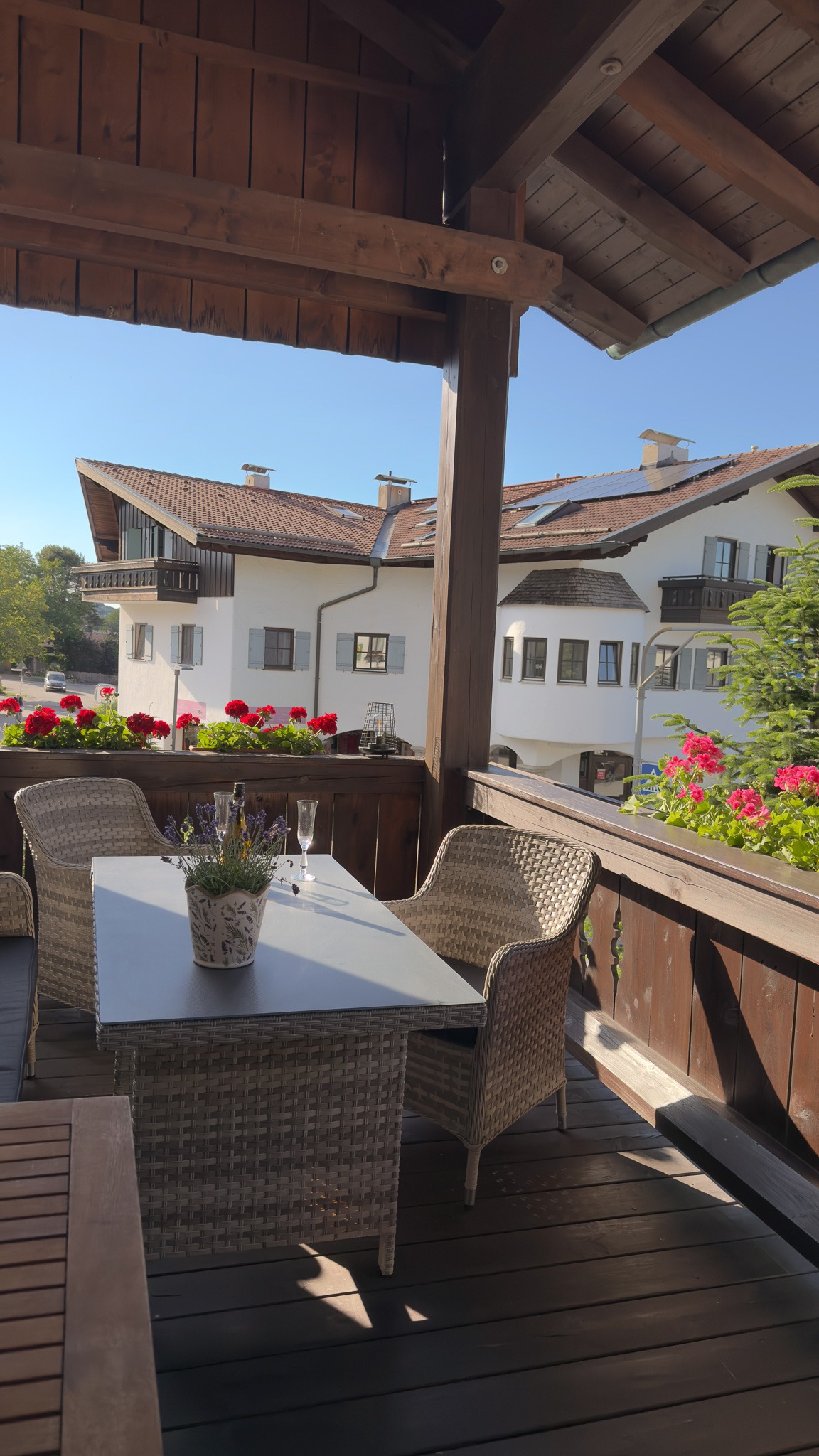 Balkon mit komfortablen Rattanmöbeln, roten Geranien am Geländer und einem schönen Blick auf traditionelle bayerische Häuser in der Nachbarschaft.