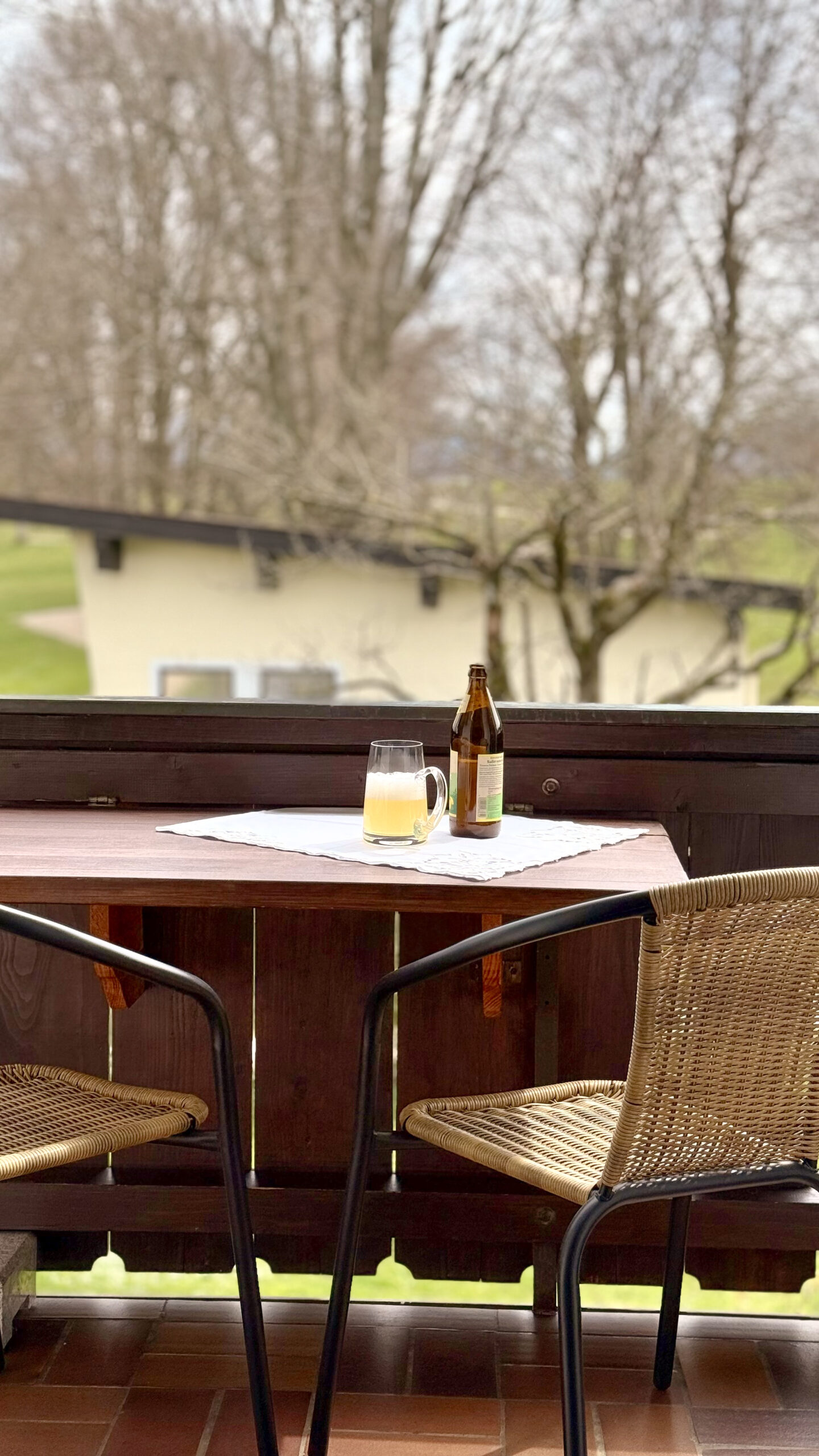 Ein Holztisch auf einem Balkon mit zwei Korbstühlen. Auf dem Tisch stehen ein eingeschenktes Glas und eine Flasche Bier, im Hintergrund sieht man grüne Natur.