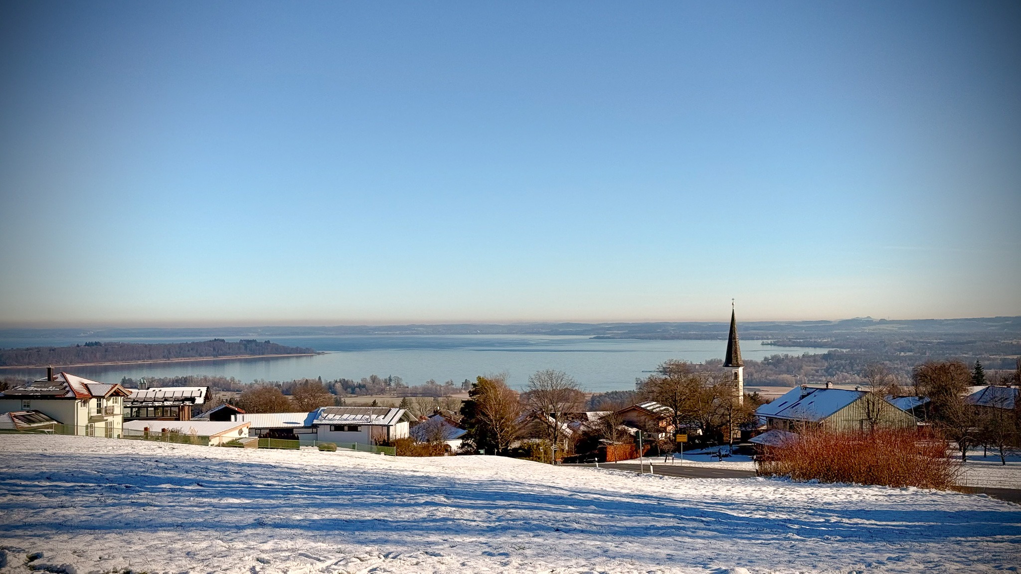 Blick über das verschneite Dorf Bernau am Chiemsee mit seinem markanten Kirchturm, dahinter der weite Chiemsee im Winter.