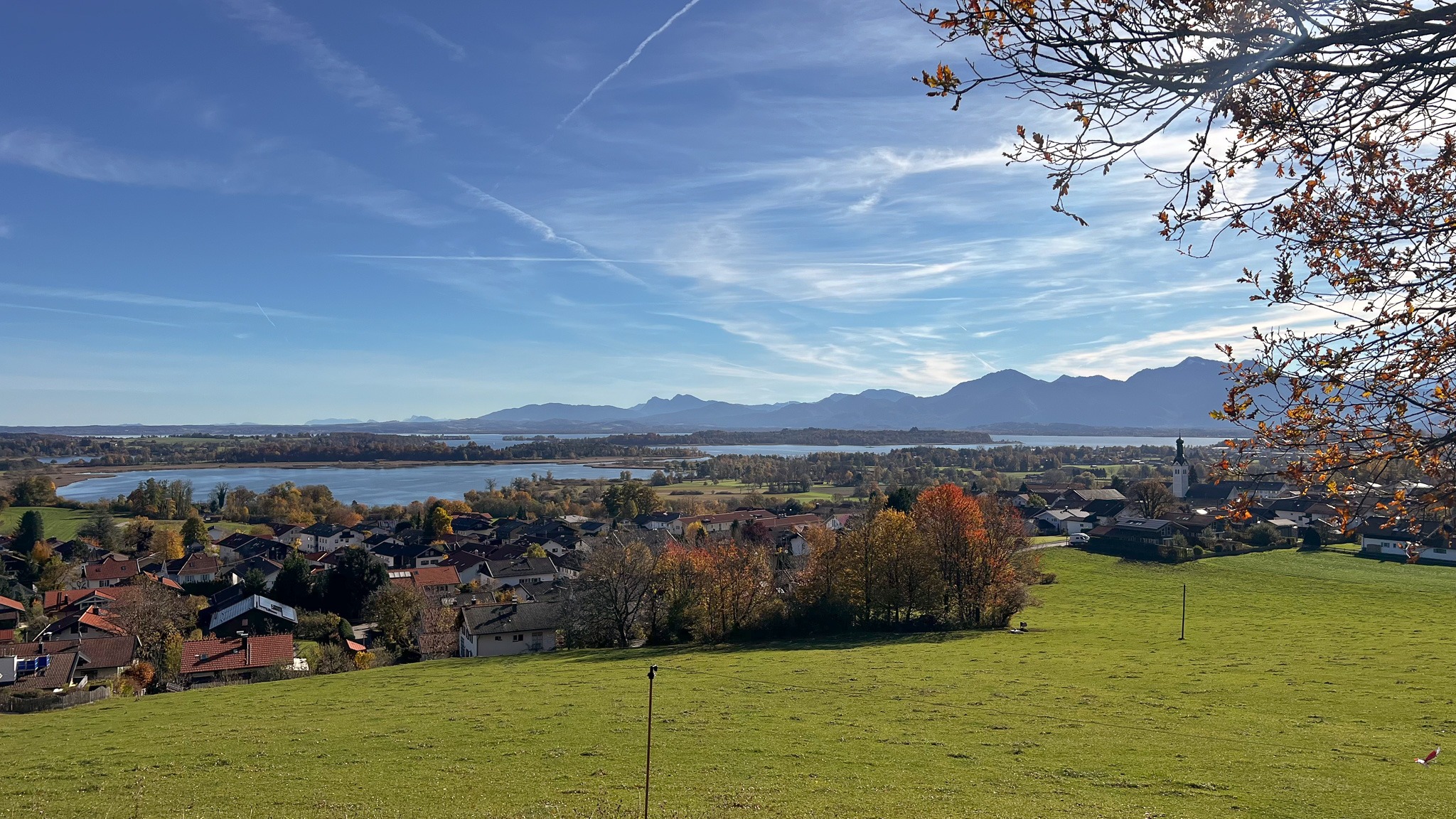 Weite Landschaftsaufnahme im Chiemgau mit grünen Wiesen, einem malerischen Dorf, dem Chiemsee und den Bergen im Hintergrund.