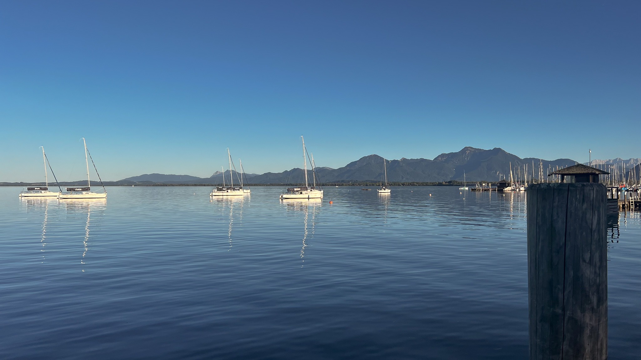Ruhiger Chiemsee am Morgen mit weißen Segelbooten auf dem Wasser und der beeindruckenden Chiemgauer Alpenkette im Hintergrund.