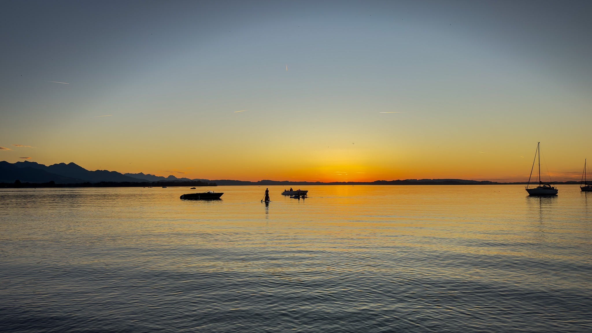 Romantischer Sonnenuntergang am Chiemsee. Die goldenen Sonnenstrahlen spiegeln sich im Wasser, während Stand-Up-Paddler und kleine Boote als Silhouetten zu sehen sind.