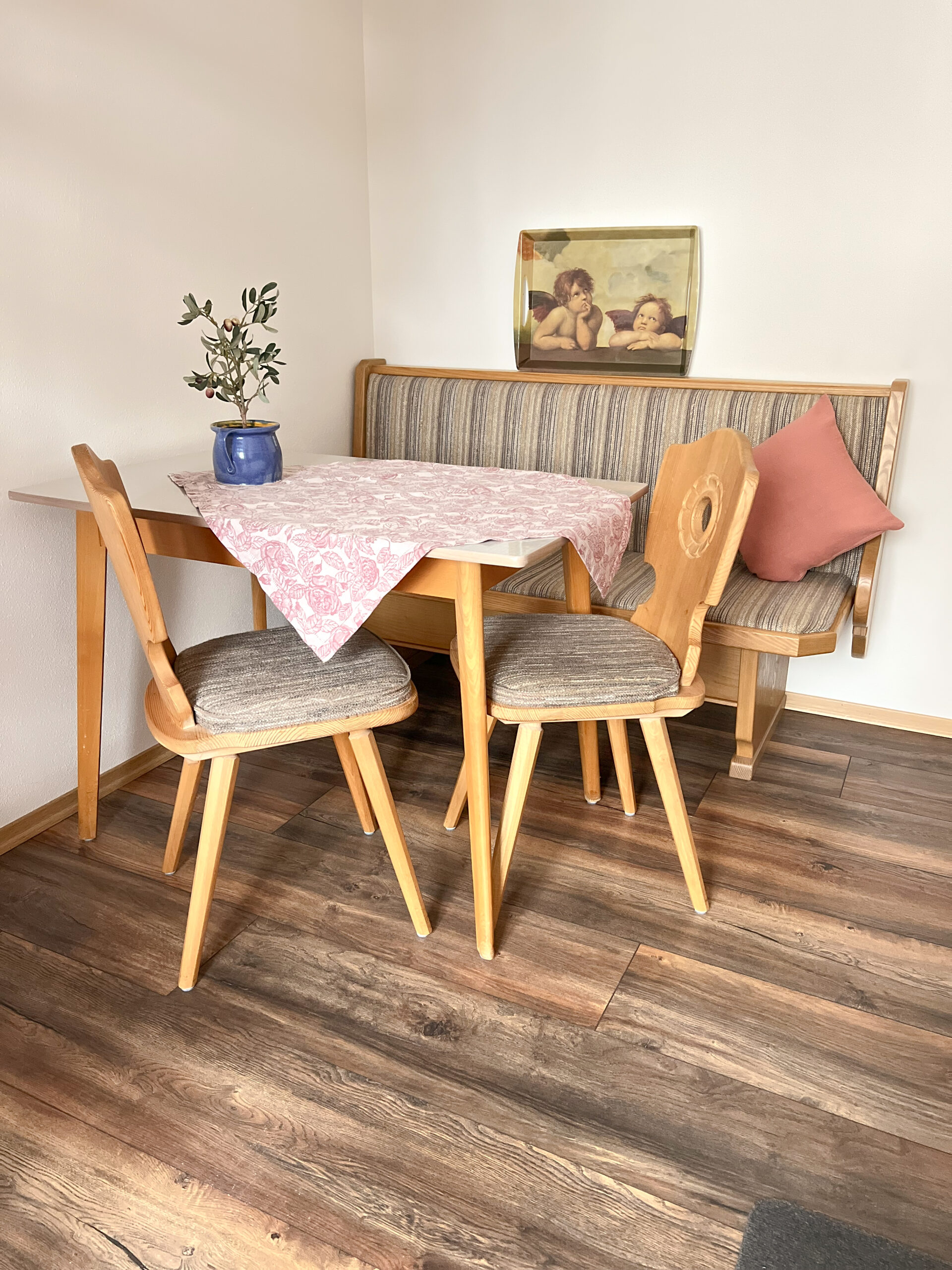 Ein gemütlicher kleiner Essbereich mit einer holzvertäfelten Eckbank, zwei Bauernstühlen und einem Tisch mit rosafarbener Tischdecke, darauf steht eine kleine Olivenpflanze.