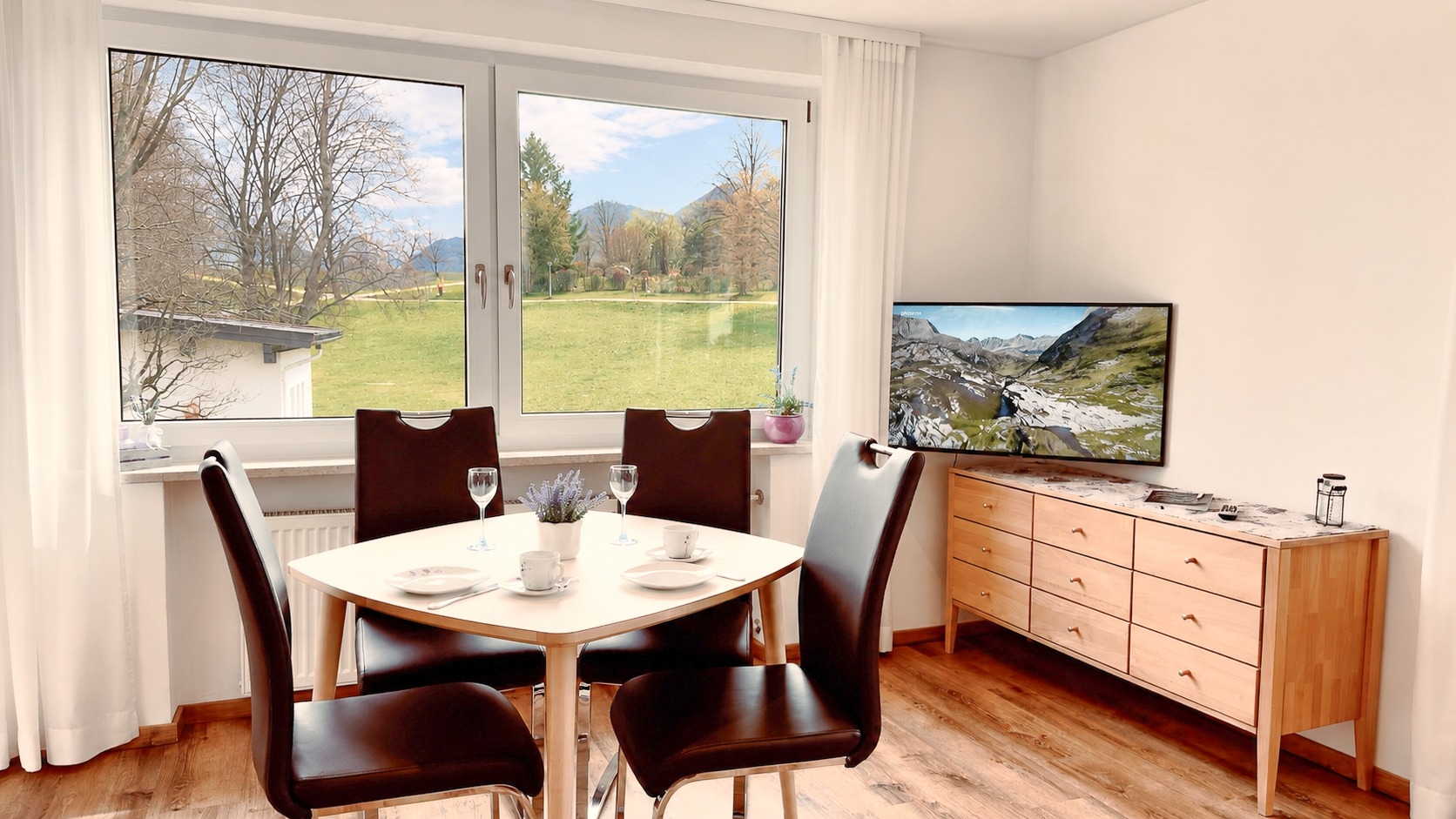 Ein heller Essplatz mit Holztisch und vier schwarzen Stühlen direkt vor großen Fenstern mit weitem Blick auf die Wiesen und die Alpen.