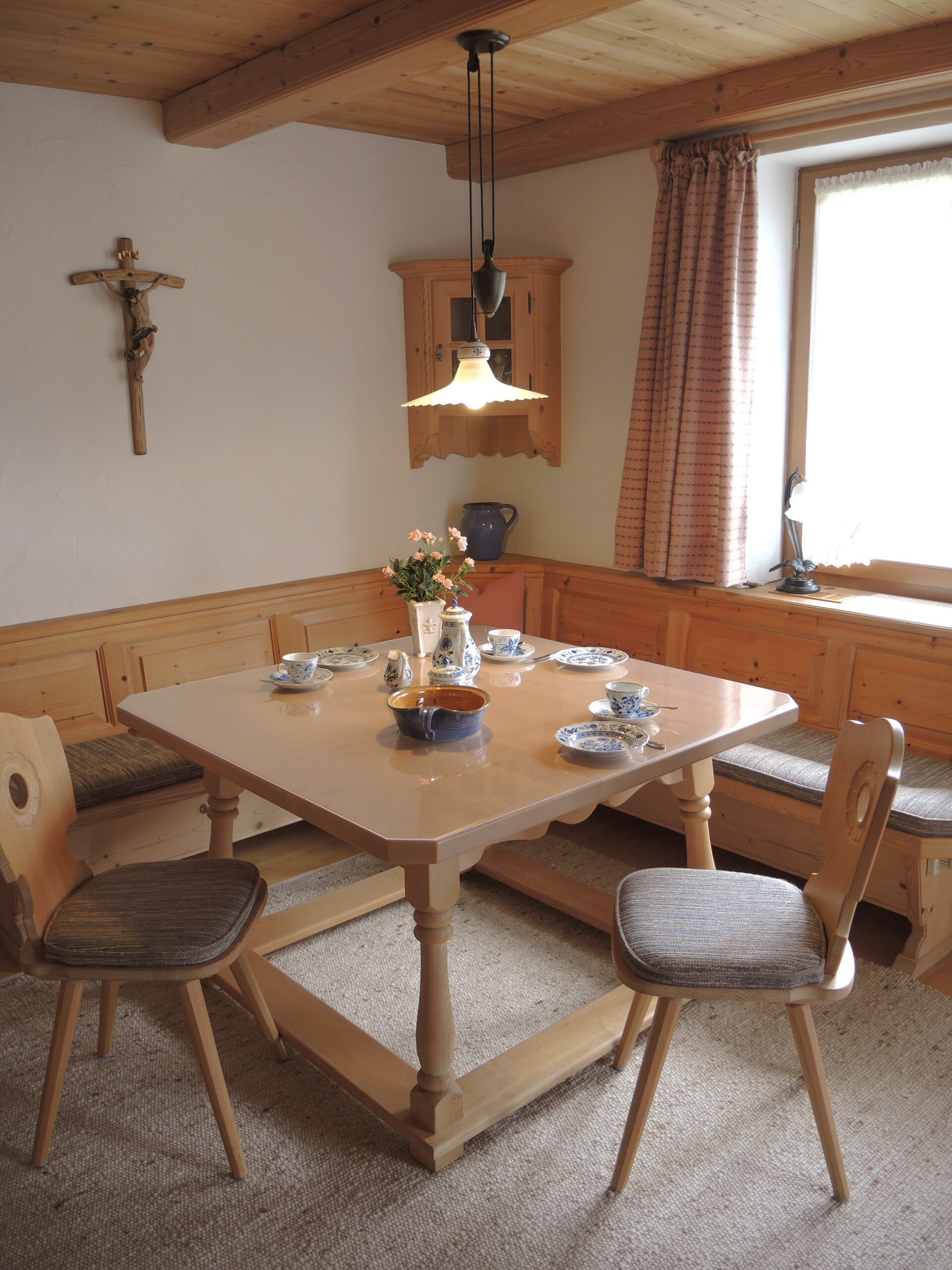 Ein traditionell bayerisches Esszimmer mit einem massiven Holztisch, einer großen Eckbank, zwei Bauernstühlen und einem gedeckten Kaffeeservice in Blau-Weiß.