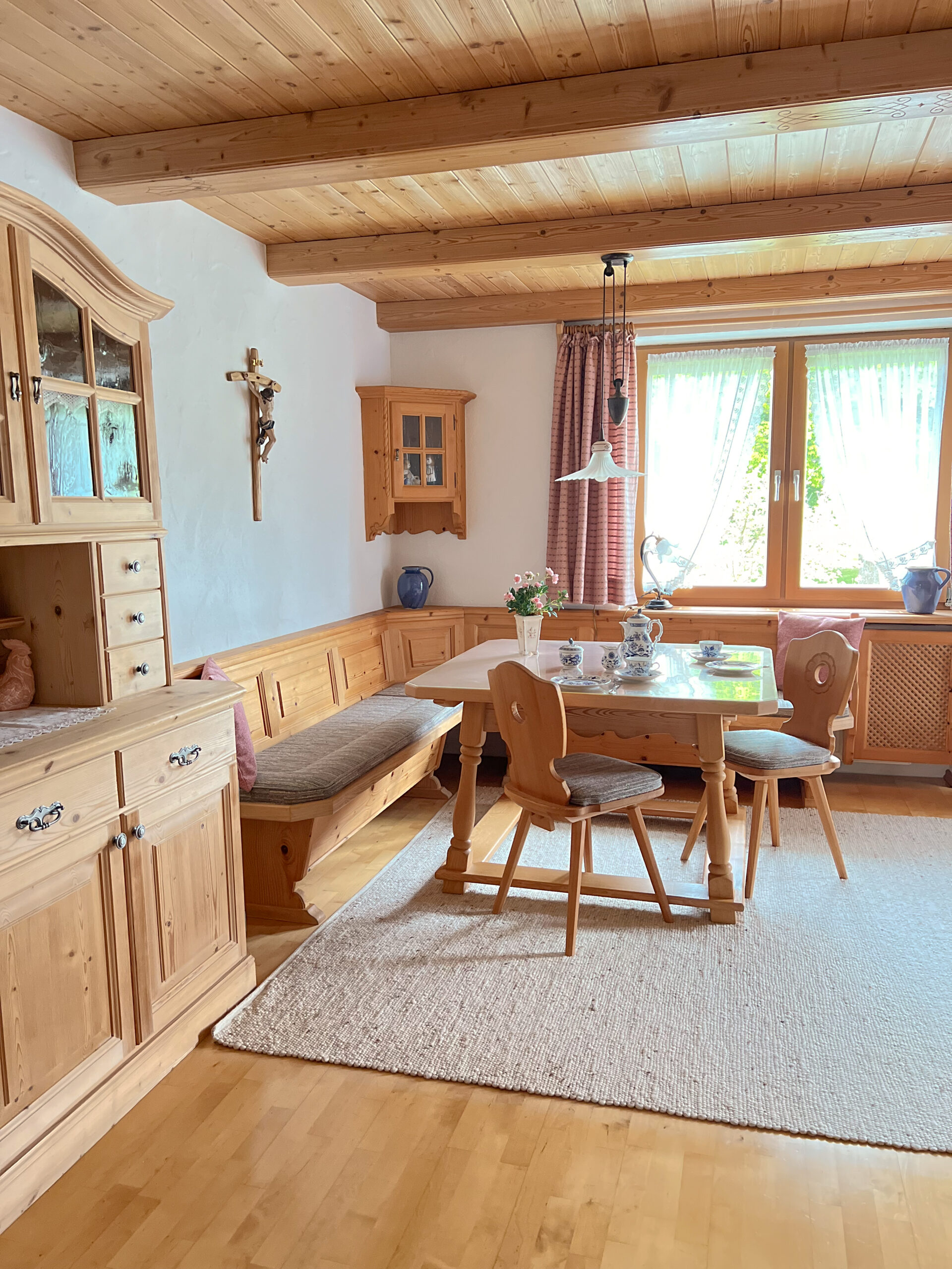 Ein traditionelles bayerisches Esszimmer mit Holzdecke, einer großen hölzernen Eckbank, einem Esstisch, einem reich verzierten Holzschrank und fein gedecktem blau-weißem Kaffeeservice.