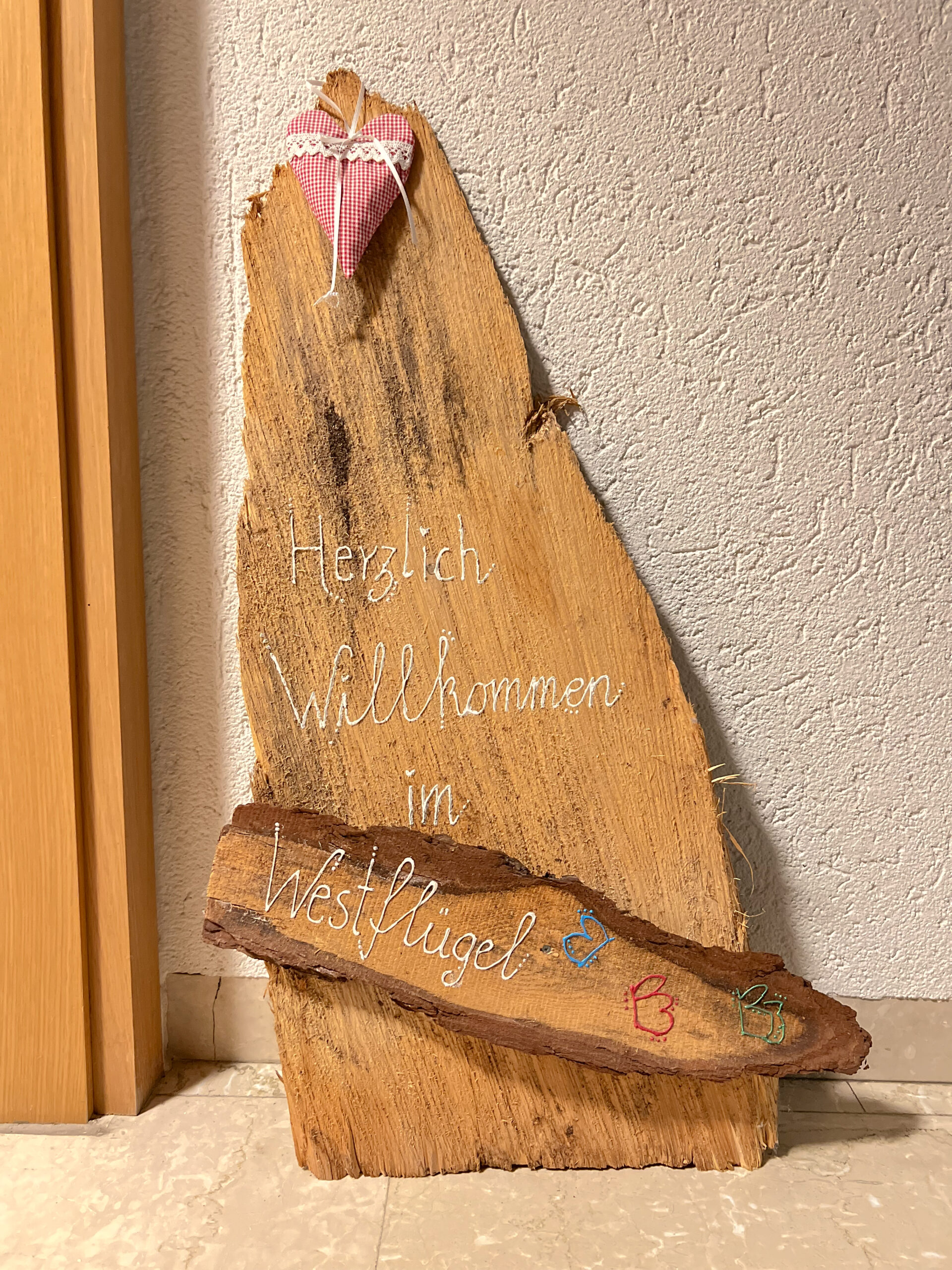 Ein rustikales Holzschild mit der weißen Aufschrift "Herzlich Willkommen im Westflügel", verziert mit einem kleinen karierten Stoffherz, steht auf dem Boden und lehnt an einer hellen Wand.