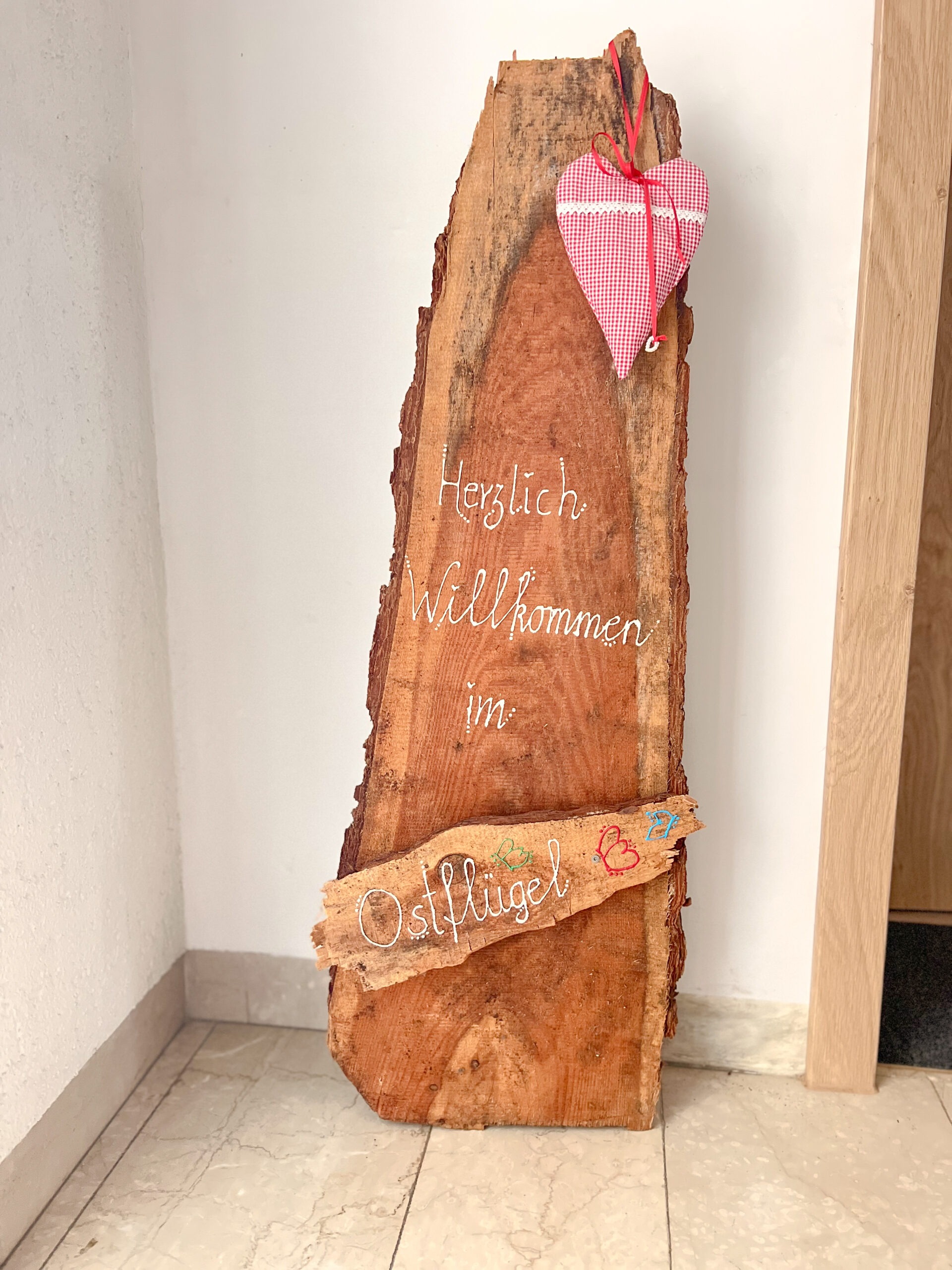 Ein hohes, rustikales Holzbrett mit der handgeschriebenen weißen Aufschrift "Herzlich Willkommen im Ostflügel", liebevoll dekoriert mit einem rot-weiß karierten Stoffherz.