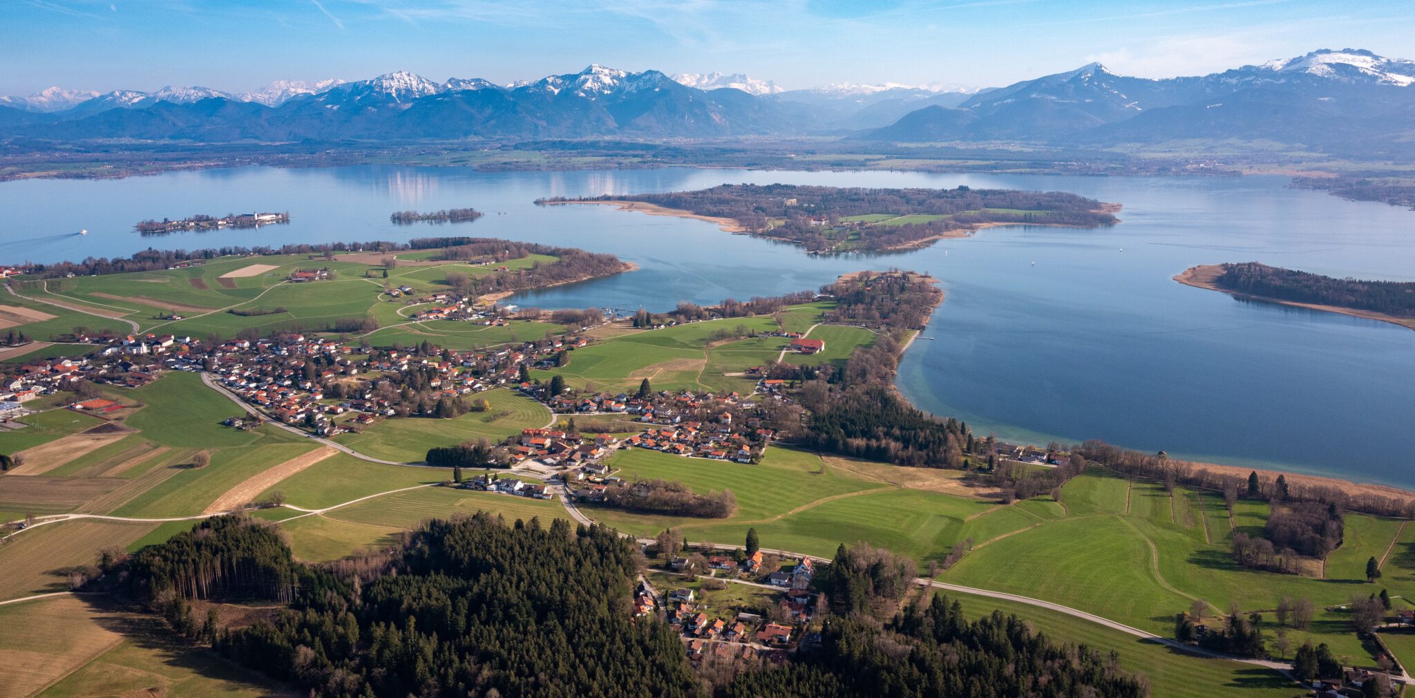 Luftaufnahme vom Chiemsee mit der Herreninsel, der Fraueninsel und den Alpen im Hintergrund, perfekt für die Planung vom Schloss Herrenchiemsee Ausflug.