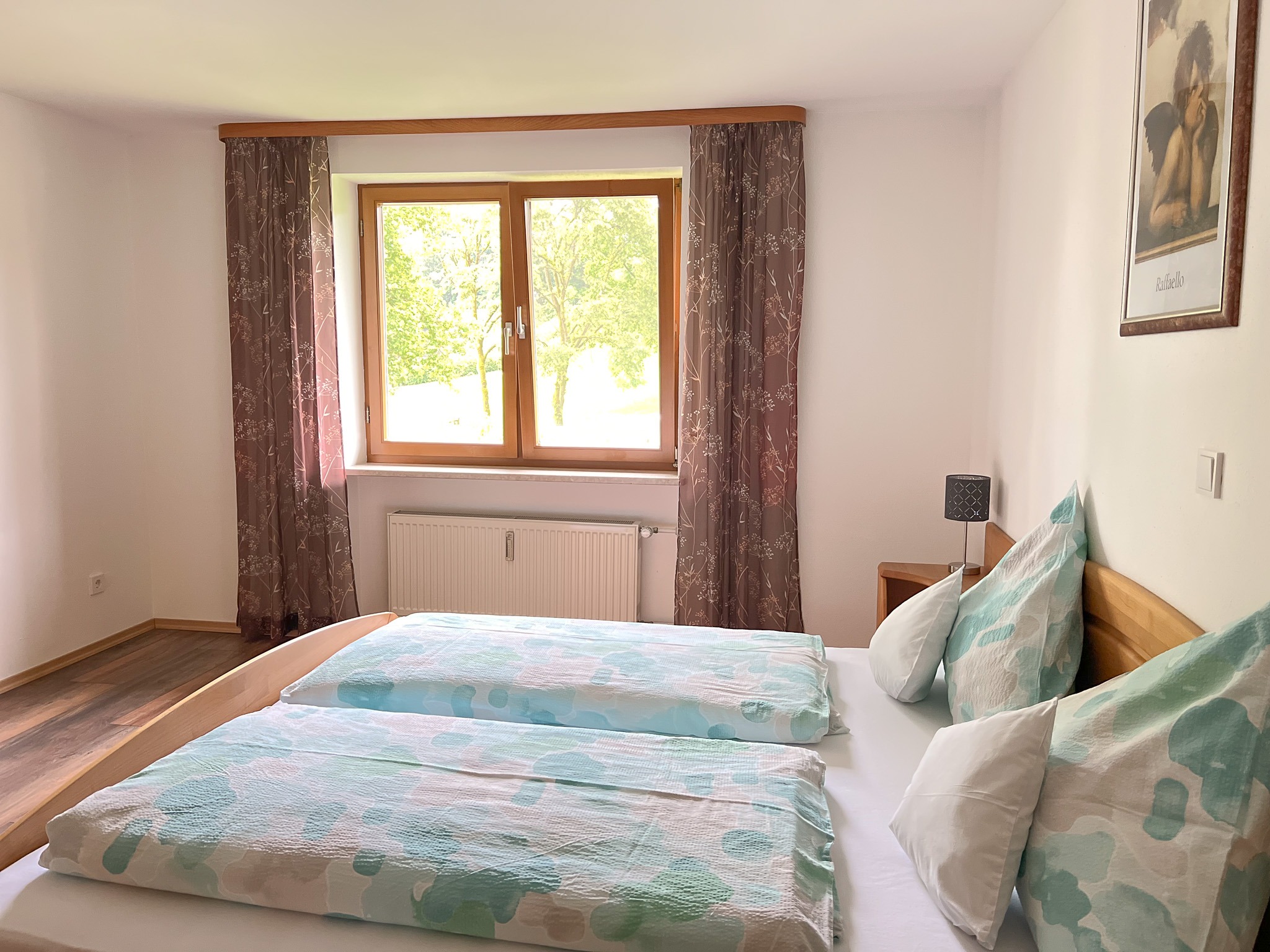 Gemütliches Doppelbett neben einem Fenster mit herrlichem Blick nach draußen ins Grüne auf sonnige Bäume.