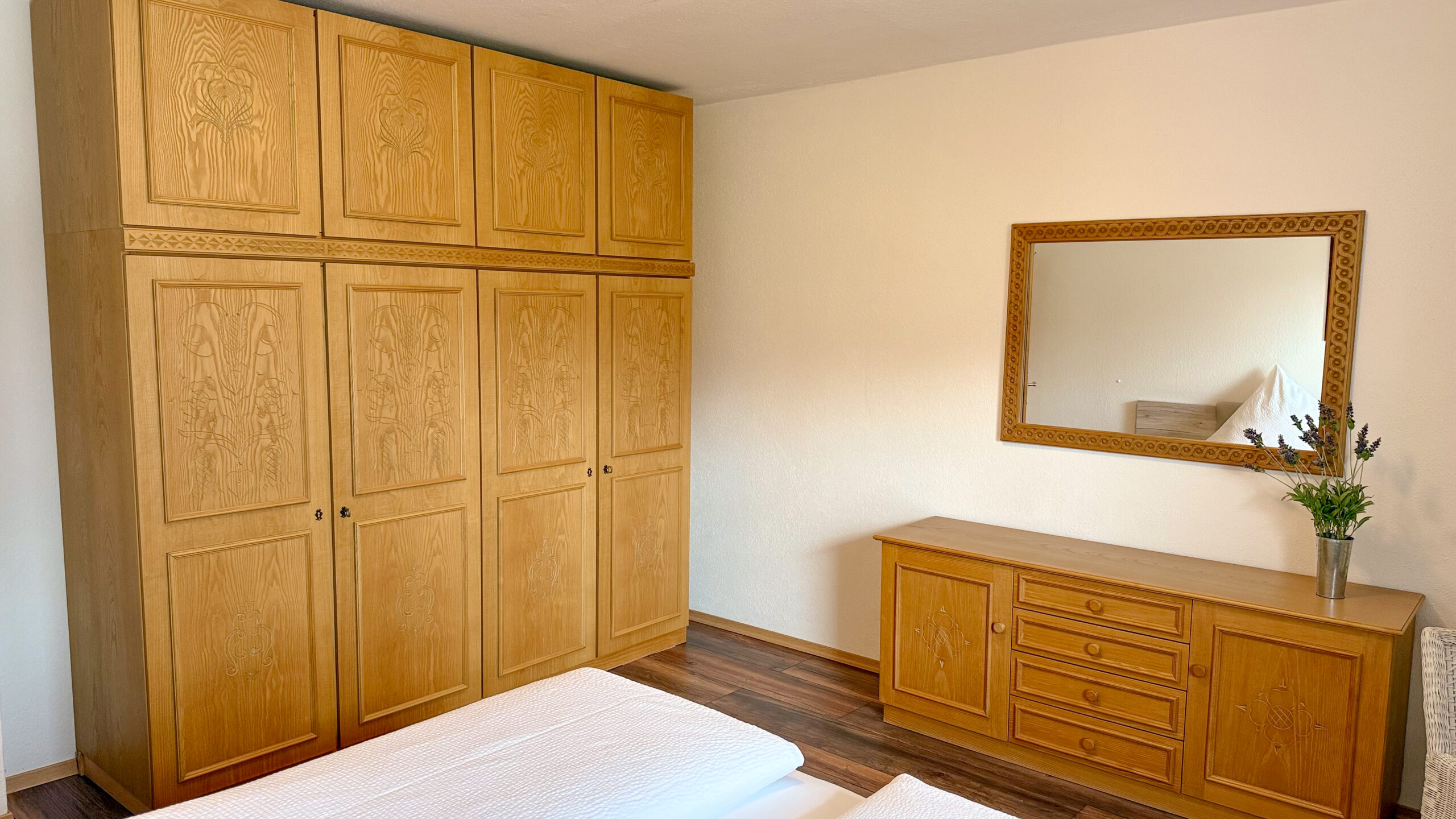 Ein massiver, deckenhoher Kleiderschrank aus Holz mit aufwendigen Schnitzereien neben einer breiten Holzkommode im Schlafzimmer.