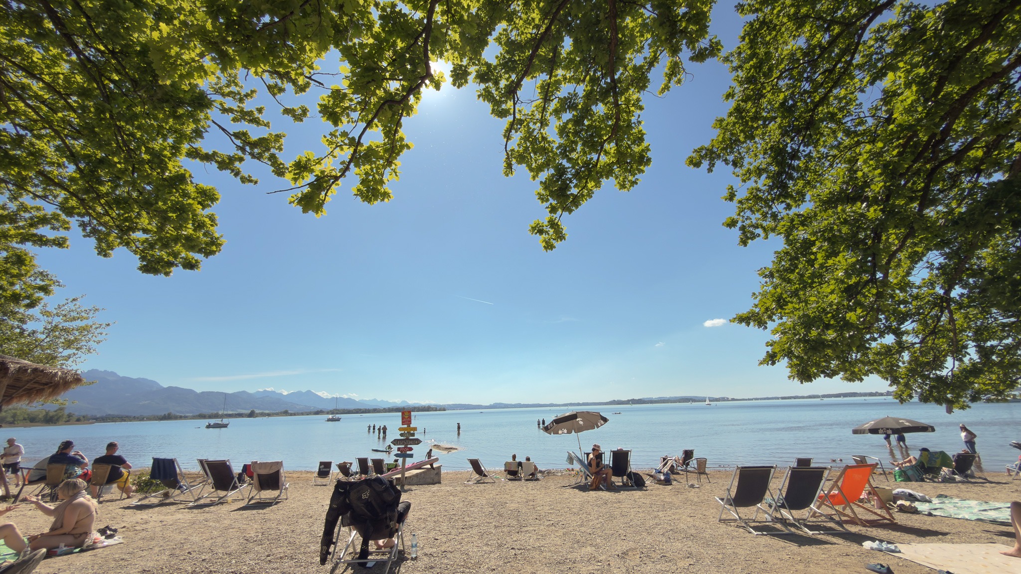 Ein idyllischer Badestrand am Chiemsee, eingerahmt von schattenspendenden Bäumen. Urlauber entspannen auf Liegestühlen im Sand, mit Blick auf das Wasser und die Alpen.