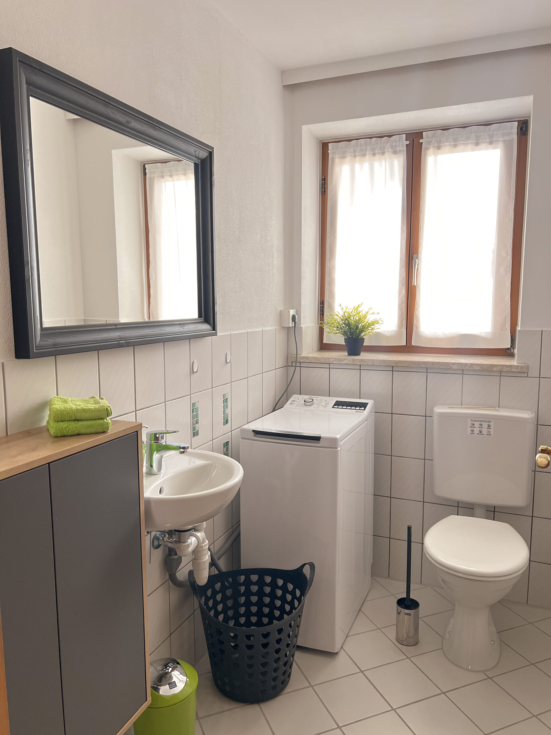 Ein helles Badezimmer mit Holzfenster, ausgestattet mit einer Waschmaschine, einem WC, einem kleinen Handwaschbecken und einem Spiegel mit dunklem Rahmen.