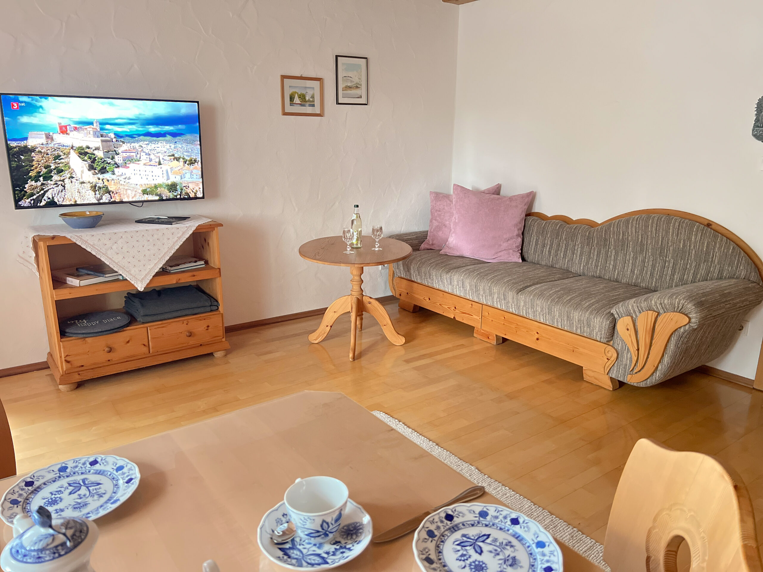 Ein gemütliches Wohnzimmer mit einem traditionellen Sofa, einem Flachbildfernseher auf einem Holzschrank und einem Esstisch im Vordergrund, der mit blau-weißem Porzellan gedeckt ist.
