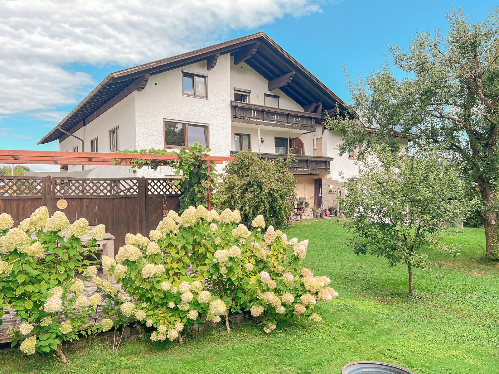 Ferienwohnung am Chiemsee: Hausansicht mit Holzbalkonen, Pergola, Terrasse und blühenden Hortensien im Garten